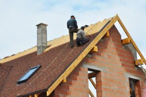 A roofing contractor and a worker install a roof for a house under construction.