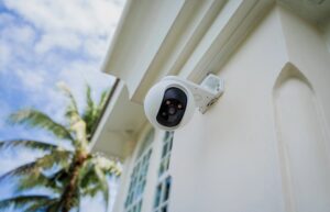 An outdoor home security camera mounted on a white wall under eaves, with a palm tree and blue sky in the background.